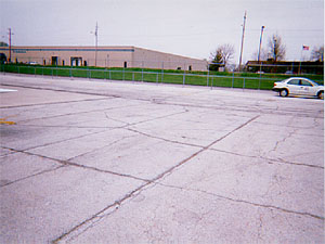 Overview photo of an apron pavement with a uniform rectangular pattern of medium-severity           tranvserse and longitudinal cracks.  The pattern is the result of cracks that have reflected through the asphalt surface at the joints of the PCC slabs below.  Some spalling is visible at the cracks.