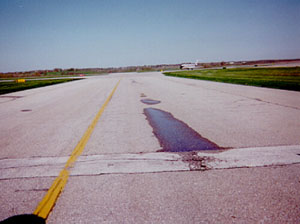 Overview photo of a taxiway pavement with visible low-severity rutting in one wheelpath.            The rutting is visible because there is standing water in the wheelpath.